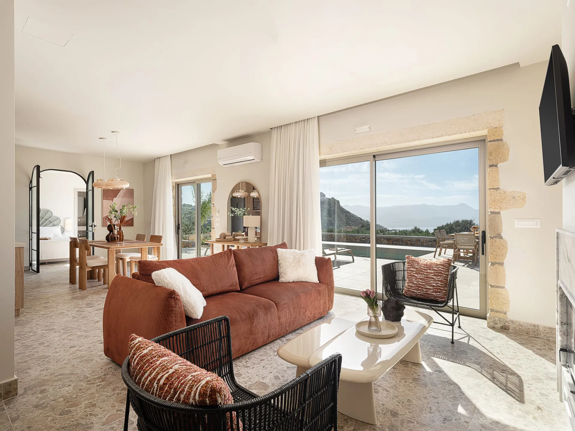 Modern open-concept living room featuring a terracotta sofa, black woven armchairs, and floor-to-ceiling glass doors with a scenic nature view.